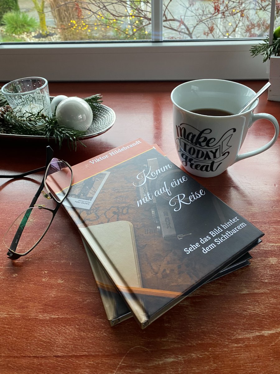 Das Buch „Komm mit auf eine Reise“ auf einem Tisch am Fenster, daneben eine Brille und eine Tasse Kaffee